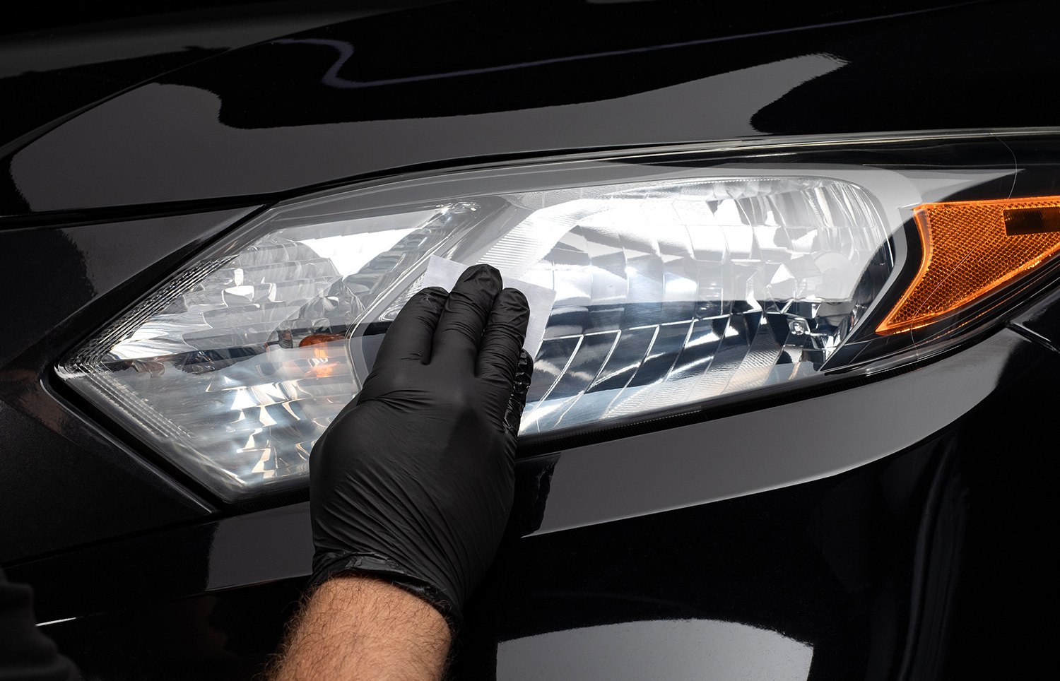 A gloved hand applying the CERAKOTE ceramic coating wipe across a clear headlight lens on a black car