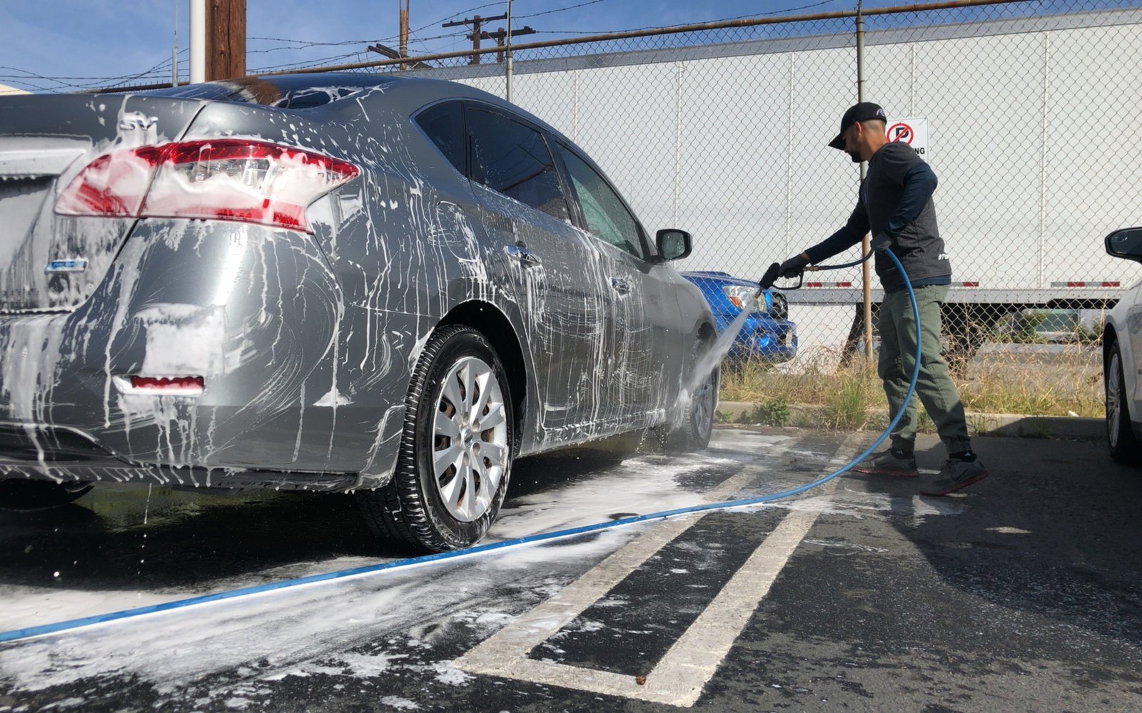A gray sedan covered in white foam being rinsed with a hose on a driveway